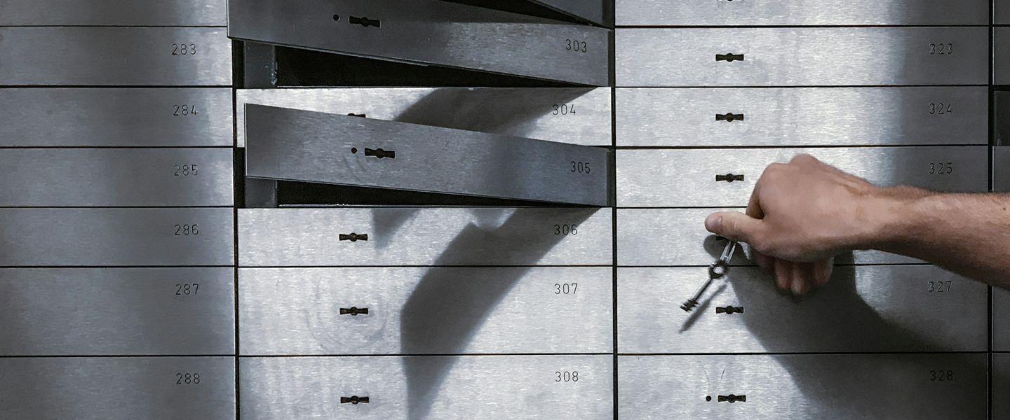 A hand holding a key in front of numbered steel safe-deposit boxes