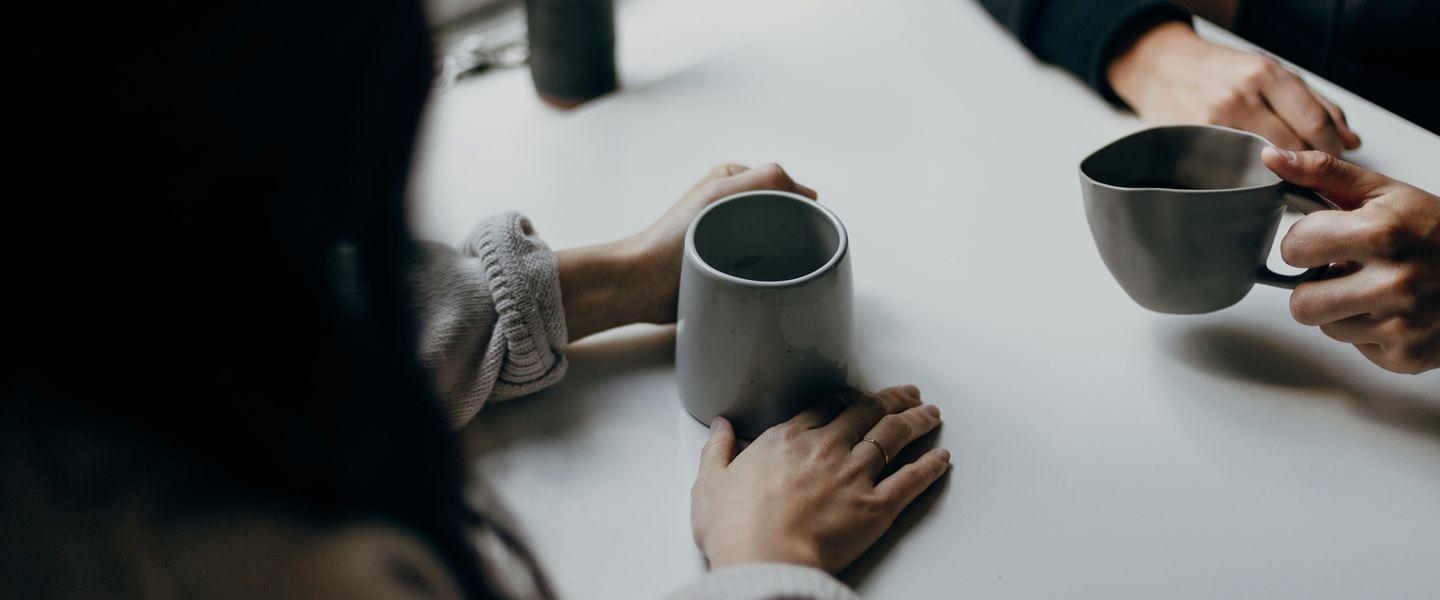 Two people talking across a white table with coffee mugs by a window