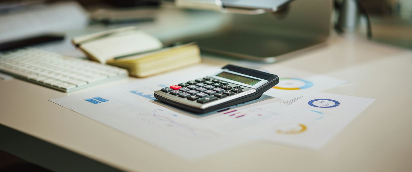 Calculator resting on printed charts on a desk, laptop blurred behind