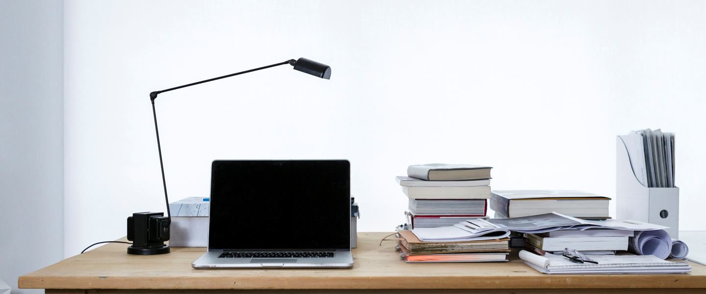 Laptop, reading lamp and stacks of books on a desk against a white wall